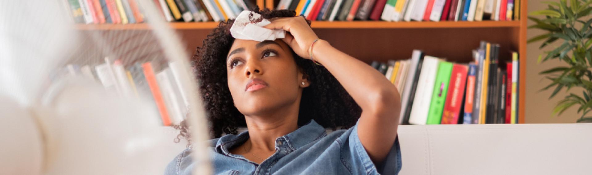 Woman Warm In Room Sitting In Front Of Fan Holding Cold Wash Cloth On Her Forehead